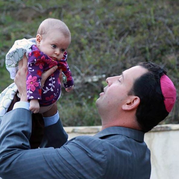 Rabbi Spike Anderson is holding up a newborn baby in a naming ceremony. The baby is wearing a pink outfit and the rabbi is wearing a pink kippah.