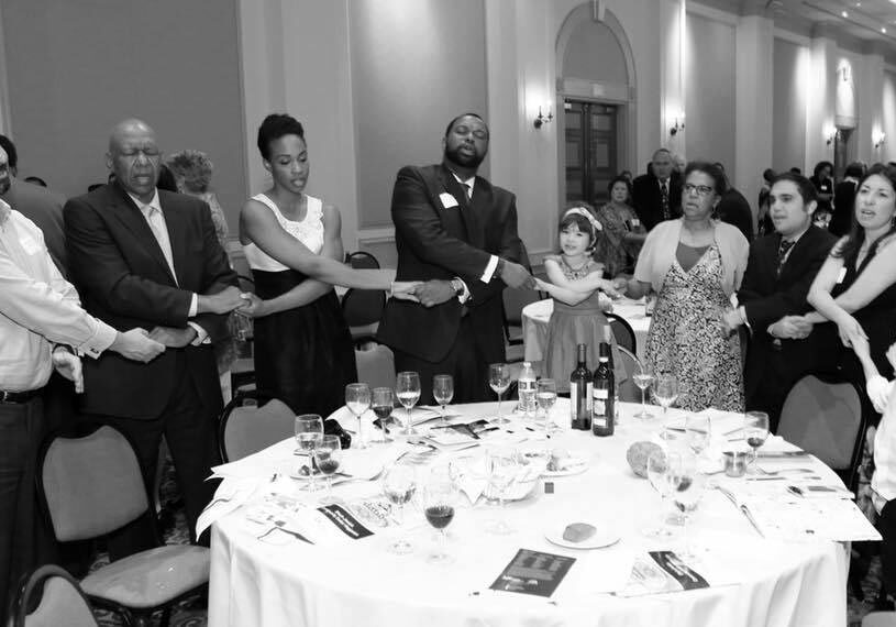 Rabbi Spike and family at the Black Jewish Seder meal at The Temple. A group of attendees, both black and white, are holding hands in prayer and song around the seder table. The image is black and white.