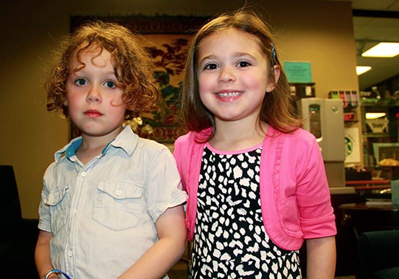 Two little girls standing together and smiling. One is wearing a button down shirt and the other is wearing a black and white dress with a pink cardigan.
